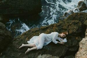 sensueel vrouw in lang wit jurk nat haar- aan het liegen Aan een rotsachtig klif landschap foto