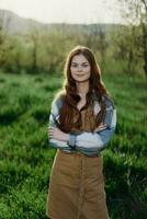 een boer vrouw in haar werk kleren, plaid overhemd en schort, staat in de veld- Aan de groen gras en glimlacht in de instelling zon foto