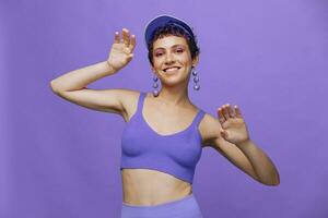 sport- mode vrouw dansen poseren glimlachen met tanden in een Purper sport- pak voor yoga Aan een slank lichaam en een transparant pet Aan haar hoofd Aan een Purper achtergrond monochroom foto