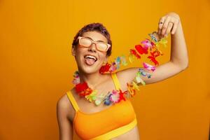 portret van een vrouw met een bloemen hawaiiaans krans in de omgeving van haar nek is hebben pret dansen en glimlachen in helder kleren Aan een oranje achtergrond vervelend zonnebril, levensstijl partij hawaiiaans stijl foto