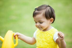 zoet een jaar oud baby meisje gekleed in geel gieter de planten Bij de tuin foto