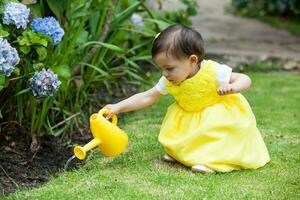 zoet een jaar oud baby meisje gekleed in geel gieter de planten Bij de tuin foto