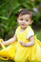 zoet een jaar oud baby meisje gekleed in geel gieter de planten Bij de tuin foto