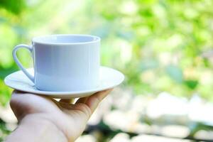 hand- Holding wit keramisch kop van heet koffie met natuurlijk licht ochtend- in de tuin. cafeïne drankjes oorzaak een vers gevoel. foto