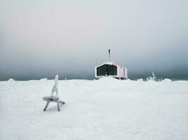 selectief focus. houten bank tegen de achtergrond van een eenzaam rood gast huis Aan een met sneeuw bedekt helling in winter onder dramatisch lucht. dubbele huis Aan de berg volodyanaja kandalaksha in Rusland. foto