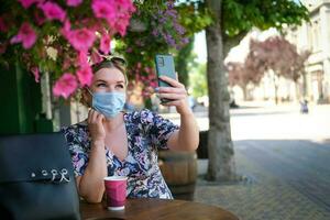 mooi jong vrouw in een cafe in een medisch masker en maakt een selfie Aan de telefoon. een meisje in een cafe Aan de straat communiceert online Aan een smartphone. pandemisch covid19, quarantaine. foto