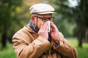 buitenshuis portret van een senior Mens in winter jas blazen zijn neus- foto