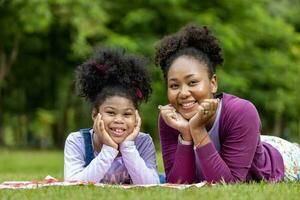 Afrikaanse Amerikaans moeder en jong dochter zijn aan het liegen naar beneden na hebben een zomer picknick in de openbaar park voor weekend vrije tijd en geluk concept foto