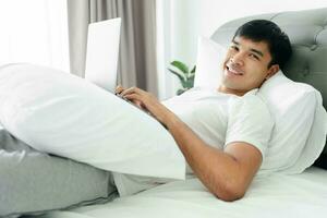 Aziatisch Mens in wit t-shirt houdende Aan bed gebruik makend van laptop computer in slaapkamer. foto