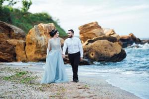 bruid in een blauwe jurk met bruidegom wandelen langs de oever van de oceaan foto