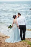 bruid in een blauwe jurk met bruidegom wandelen langs de oever van de oceaan foto