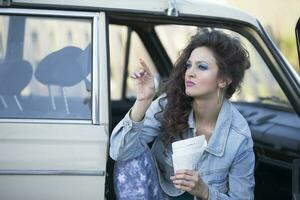 grappig jong vrouw in een denim jasje met een zak van zonnebloem zaden zit in een oud auto. vrouw in de stijl van de jaren tachtig. foto