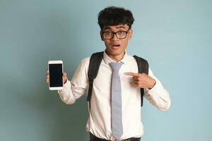 Indonesisch senior hoog school- leerling vervelend wit overhemd uniform met grijs stropdas tonen en presenteren blanco scherm mobiel telefoon terwijl richten Aan zijn smartphone. geïsoleerd beeld Aan blauw achtergrond foto