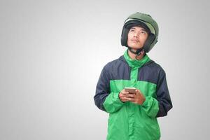 portret van verward Aziatisch online taxi bestuurder vervelend groen jasje en helm denken over een idee en op zoek omhoog. geïsoleerd beeld Aan wit achtergrond foto