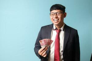 portret van jong Aziatisch bedrijf Mens in gewoontjes pak lachend uit luid Holding duizenden roepia. geïsoleerd beeld Aan blauw achtergrond foto