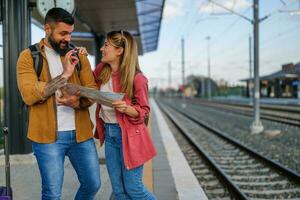 gelukkig paar is staand Bij spoorweg station en aan het wachten voor aankomst van hun trein. ze zijn op zoek Bij kaart. foto