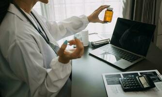 echt vrouw dokter gebruik makend van laptop en schrijven aantekeningen in medisch logboek zittend Bij bureau. jong vrouw professioneel medisch arts vervelend wit jas en stethoscoop werken Aan computer Bij werkplek. foto