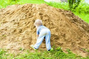 een weinig jongen klimt een berg met zand. de kind is allemaal vies, maar gelukkig. foto