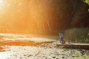 weinig jongen staand en visvangst Aan een houten dok Bij de zonsondergang foto
