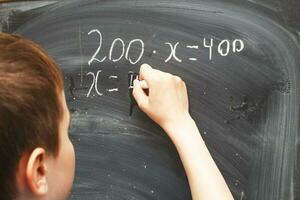 jongen staand terug in voorkant van school- schoolbord en schrijven. schooljongen lost op wiskunde voorbeeld Bij de schoolbord foto