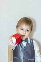 een weinig jongen houdt en handen over- een rood doos, een Valentijnsdag dag thema concept. portret van een schattig jongen in een pak met een boog binden. foto