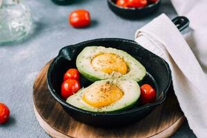 rauw avocado helften, klaar naar bakken, gevuld met ei en kers tomaten in een pan Aan de tafel. gezond aan het eten. flexitariër eetpatroon foto