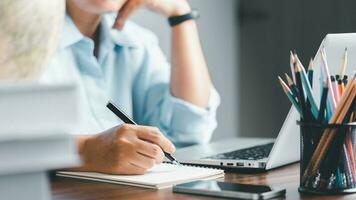 onderwijs concept. dichtbij omhoog van vrouw leerling maken aantekeningen hand- schrijven in notitieboekje studie online Aan computer van huis. Dames schrijven in kladblok pratend ver weg Aan webcam virtueel zoom telefoontje Aan laptop. foto