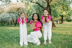 schattig mam en twee dochters blazen Aan de gedaald bloemblaadjes van appel bloesems in de voorjaar tuin. buitenshuis werkzaamheid. zoet momenten familie foto