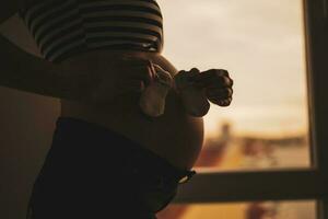 zwanger vrouw staand naast venster en Holding baby sokken. silhouet van zwanger vrouw buik. foto