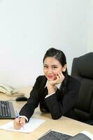 jong Aziatisch vrouw vervelend pak zittend Bij kantoor bureau kijken Bij camera hand- Aan wang foto