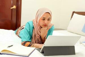 jong Aziatisch Maleis moslim vrouw vervelend hoofddoek baju kurung jurk Bij huis Aan bed studie werk Aan computer tab boek notitieboekje pen lezen schrijven foto