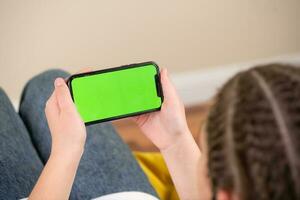 jong meisje zittend Aan een sofa en gebruik makend van een smartphone met groen scherm foto