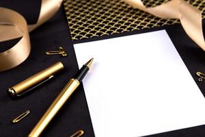 gouden pen, lint, paperclips en briefpapier op een zwarte achtergrond met een wit vel papier met kopie ruimte foto
