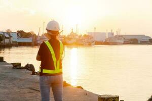 silhouetten van Aziatisch vrouw ingenieur Holding sleutels en radio communicatie ze staand Aan scheepswerf. achtergrond is olie opslagruimte silo. concept van Dames meisje macht Gelijk kans. foto