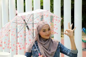 jong Aziatisch Maleis moslim vrouw hoofddoek buitenshuis groen houding met paraplu OK vinger zingen foto