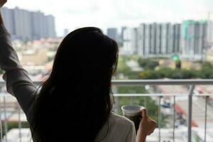 mooi jong zuidoosten Aziatisch vrouw Holding thee koffie kop in de buurt balkon venster stad scape achtergrond, op zoek ontspannende denken foto