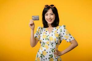 portret jong Aziatisch vrouw gelukkig glimlachen in lente jurk tonen plastic credit kaart geïsoleerd Aan geel achtergrond. betalen, geld en aankoop boodschappen doen betaling concept. foto