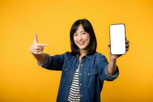 portret mooi jong Aziatisch vrouw gelukkig glimlach gekleed in denim jasje tonen smartphone scherm met richten vinger hand- gebaar naar mobiel telefoon isoleren Aan geel studio achtergrond. foto