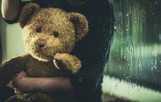 kind met teddy beer zittend Aan vensterbank Bij nacht. foto