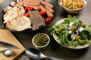 mengen ontmoeten kip rundvlees steak worst tomaat ui knoflook bakken rooster olie metaal bakken pan kleurrijk pasta groen salade met kaas olijf- munt basilicum pesto saus bord mes vork lepel rustiek houten tafel foto