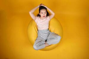 mooi jong zuiden oosten- Aziatisch vrouw zit Aan een geel zitzak stoel oranje geel kleur achtergrond houding mode stijl elegant schoonheid humeur uitdrukking rust uit kom tot rust denken emotie top visie foto