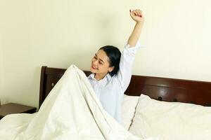 jong mooi modern Maleis vrouw kom tot rust ochtend- wakker worden omhoog Aan bed glimlach armen in lucht rekken foto