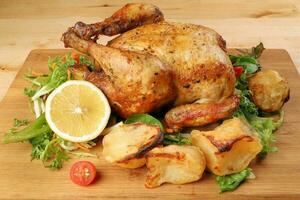 geheel geroosterd gegrild kip gevogelte vogel met gebakken aardappel groente salade tomaat citroen Aan houten snijdend bord achtergrond foto