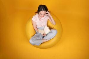 mooi jong zuiden oosten- Aziatisch vrouw zit Aan een geel zitzak stoel oranje geel kleur achtergrond houding mode stijl elegant schoonheid humeur uitdrukking rust uit kom tot rust denken emotie lezen boek top visie foto