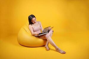 mooi jong zuiden oosten- Aziatisch vrouw zitten Aan een geel oranje zitzak stoel kleur achtergrond kom tot rust lezen studie boek tijdschrift foto