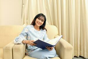 jong Aziatisch Maleis moslim vrouw vervelend baju kurung jurk Bij huis zitten rust uit Aan sofa met voeten omhoog rad boek aantekeningen kijken Bij camera foto