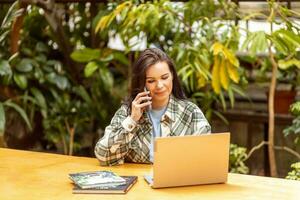 een vrouw bloemist is pratend naar klanten Aan de telefoon en plaatsen een bestellen gebruik makend van een laptop. tuin centrum, privaat bedrijf. foto