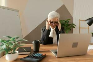 senior vrouw aanraken tempels ervaren spanning in kantoor. volwassen dame moe van werken gevoel hoofdpijn ziek wrijven tempels voorhoofd. lang laptop gebruik computer visie syndroom. kantoor syndroom. foto