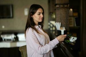 jong mooi brunette vrouw gebruik makend van contactloos betaling, vrouw klant Holding mobiel telefoon in de buurt nfc technologie Aan balie. foto