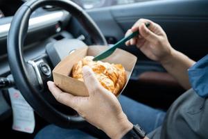 Aziatische dame die ijskoffie en broodbakkerij in auto gevaarlijk houdt en een ongeval riskeert